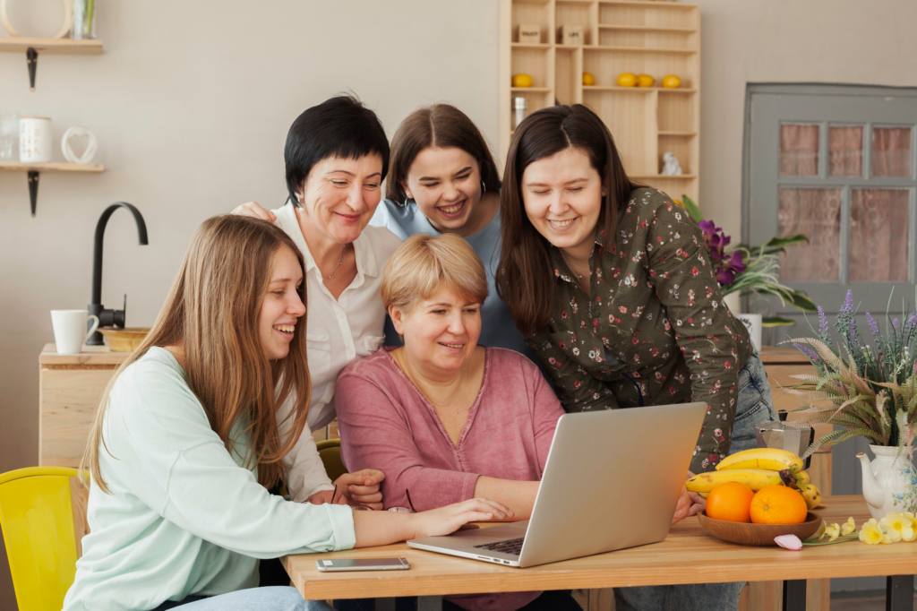 A group of five women gathered around a laptop at a kitchen table, smiling and collaborating while looking at the screen, with fruit and plants on the table.